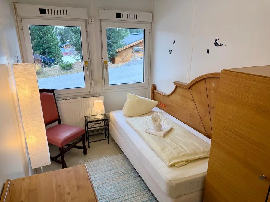 Einzelzimmer mit Bett und Holzkopfteil, rotem Stuhl, Stehlampe und Fenstern mit Blick auf Tannen