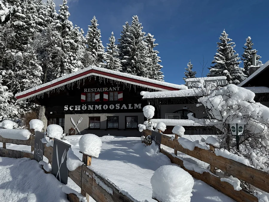 Restaurant Schönmoosalm mit verschneitem Holzzaun, Tannen und Schneehauben im Sonnenlicht