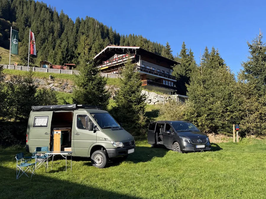 Zwei Campervans mit Campingstühlen auf grüner Wiese, Tannen und Schönmoosalm im Hintergrund