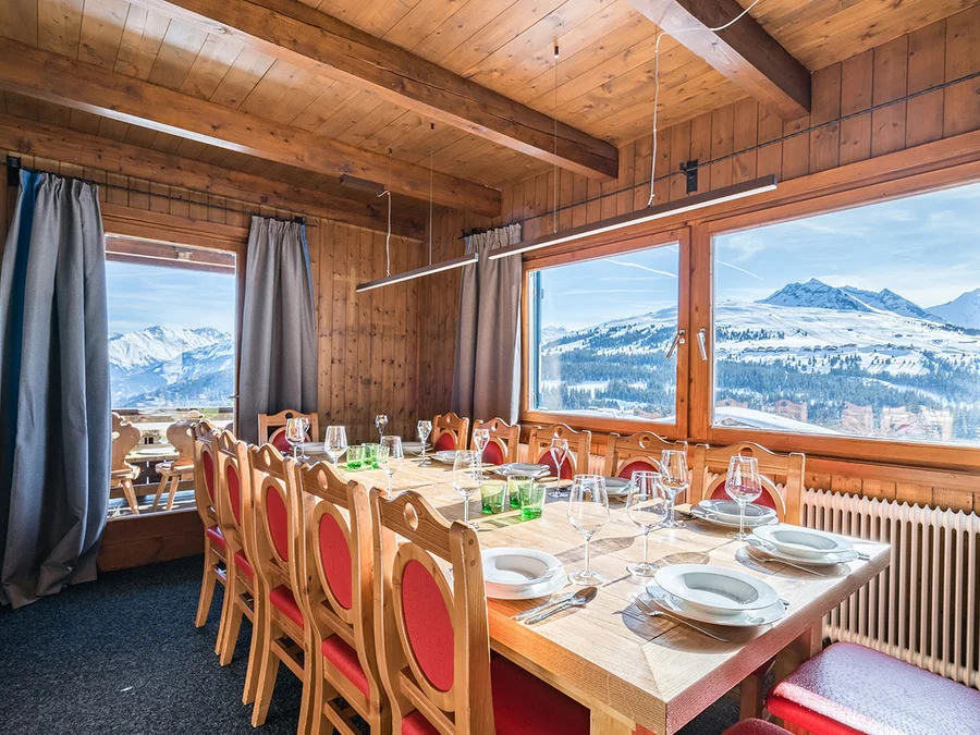 Gedeckte lange Tafel mit roten Holzstühlen im Chalet H vor Panoramafenstern mit Bergblick