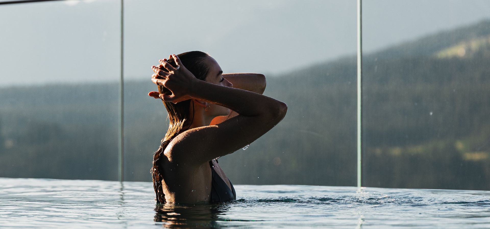 Frau im Infinity-Pool mit Glasbrüstung und Blick auf bewaldete Berge