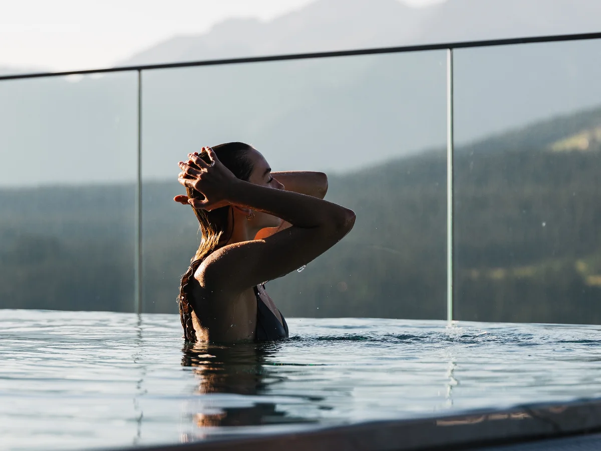 Frau im Infinity-Pool mit Glasbrüstung und Blick auf bewaldete Berge