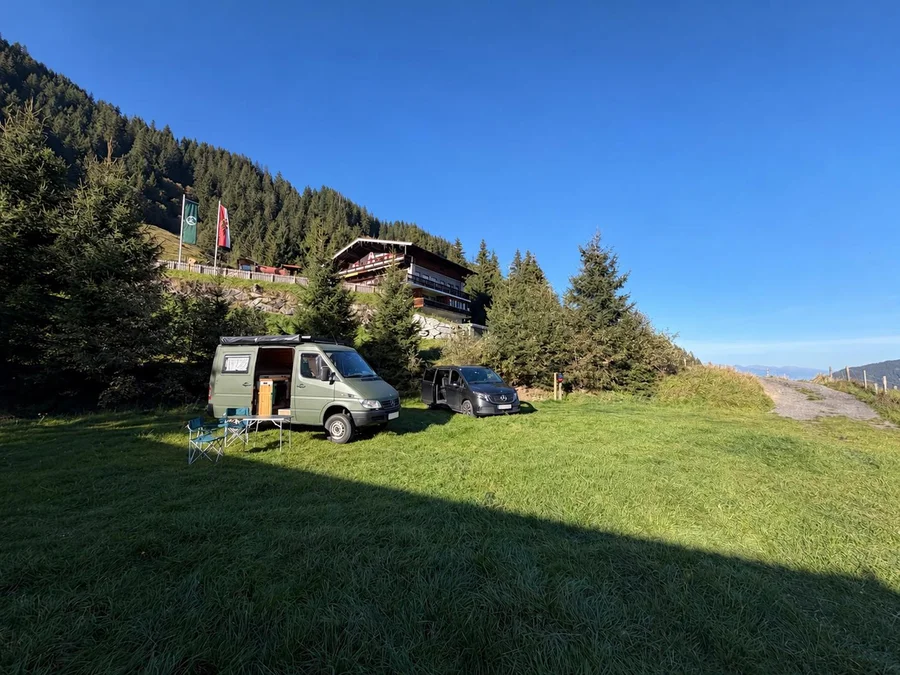 Weite Almwiese mit zwei Campervans, Tannen und der Schönmoosalm unter blauem Himmel