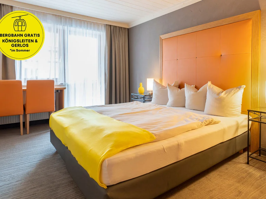 Doppelzimmer mit gelber Tagesdecke, orangefarbenem Kopfteil, zwei Stühlen und Fenster mit Vorhängen